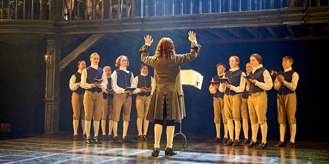A choir stand on stage, wearing smart blue waistocats, brown shorts and white tights and neck ruffs. They are all smiling and singing, reading from small black books in their hands. They look towards a conductor who has his back to the camera. He has long curly hair and wears a smart brown jacket, with his hands raised in the air.