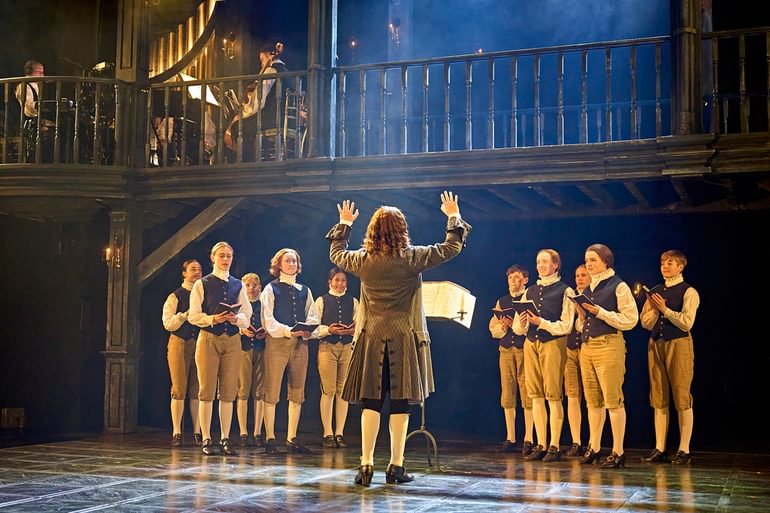 A choir stand on stage, wearing smart blue waistocats, brown shorts and white tights and neck ruffs. They are all smiling and singing, reading from small black books in their hands. They look towards a conductor who has his back to the camera. He has long curly hair and wears a smart brown jacket, with his hands raised in the air.