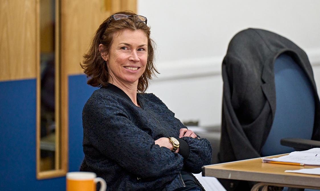 A brown-haired woman sits at a table, smiling at something out of frame. She has glasses on her head, is wearing a gold watch and has her arms crossed. She is wearing a black jumper. On the table in front of her is a script, a pencil and a mug of tea.