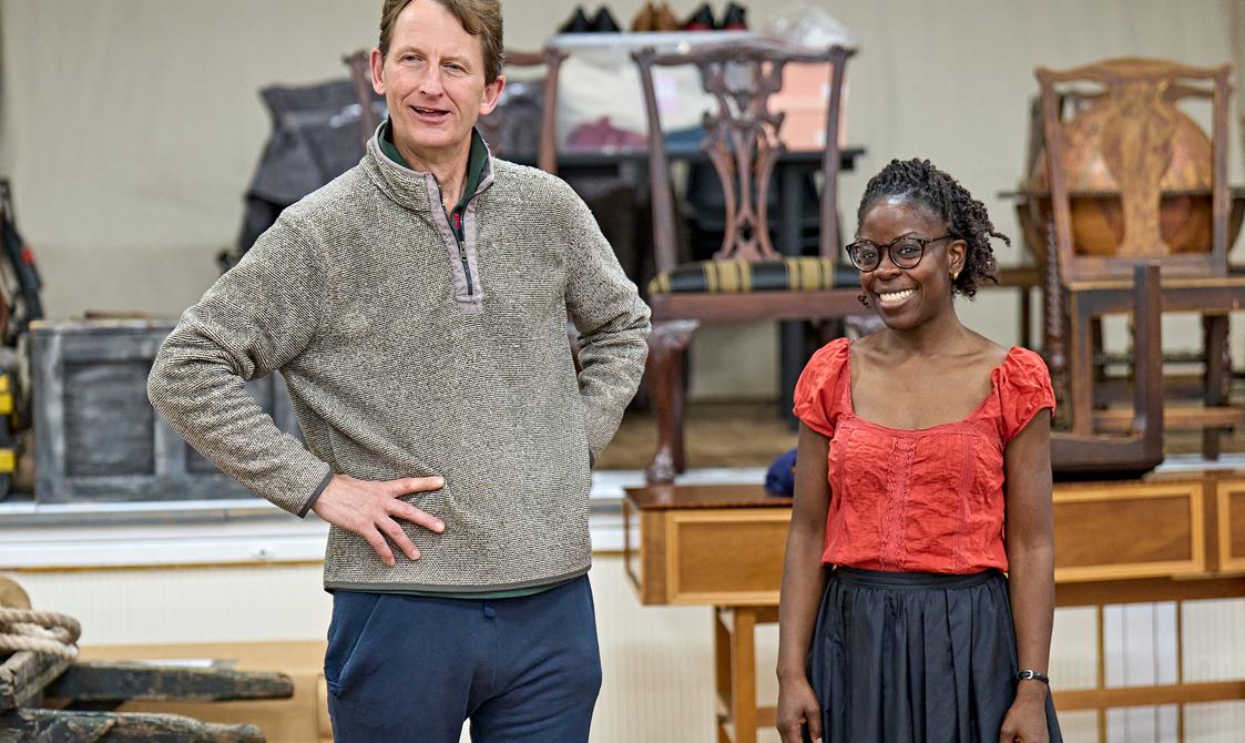 A man and a woman stand next to each other in a rehearsal room, smiling. The man has one hand on his hip and the other in his pocket, and is wearing a grey jumper and blue sweatpants. The woman has her hands casually by her sides, and wears a red blouse, a black bleated skirt, and glasses.