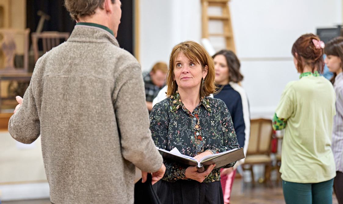 A woman in a floral shirt stands in a rehearsal room, holding a large book open in frotn of her. She has shoulder-length brown hair and is looking up at a man in front of her, who has his back to the camera. He is wearing a brown jumper, and in the background other people are stood around, talking.