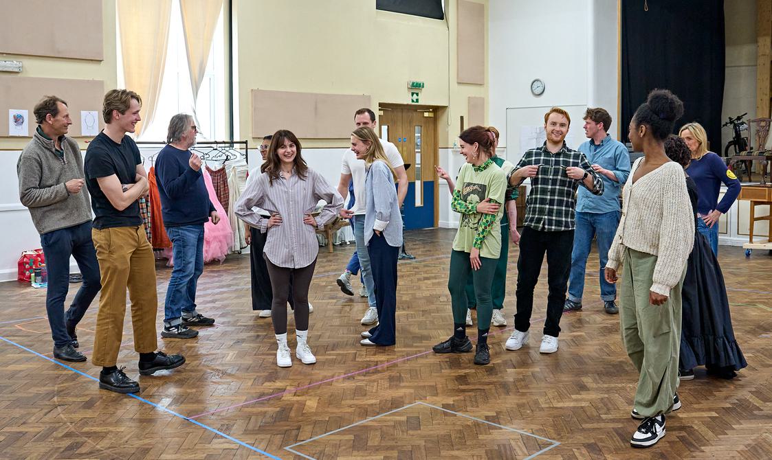 A group of actors stand around in a rehearsal room, smiling and laughing. They are all wearing different, colourful casual clothes. The floor is wooden, and in the background is a costume rail with dresses on it.