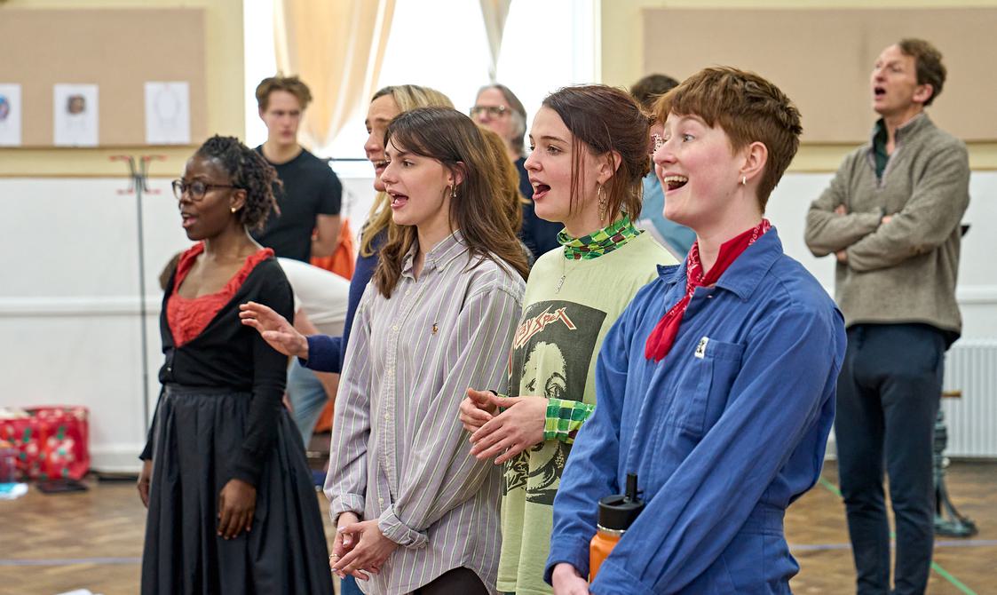 A group of actors are stood in two lines, singing. They are all facing the same direction, with their mouths open as they sing, looking happy.