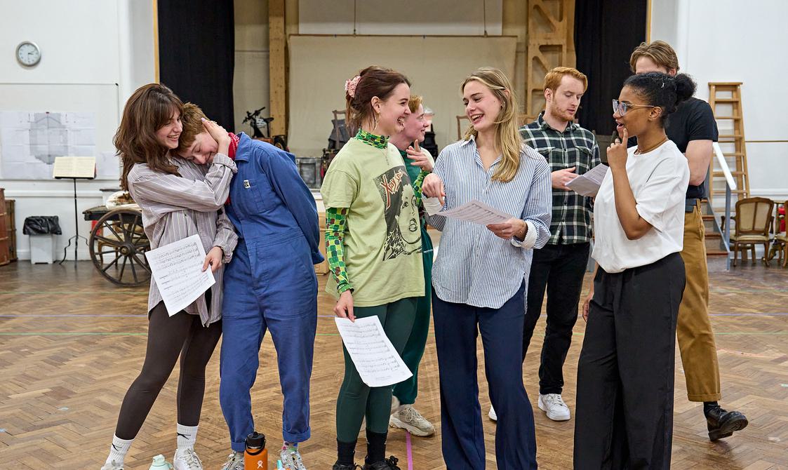 A group of actors are stood together, smiling. On the left, one has their head on another's shoulder, in the middle three people are laughing together. In the background, two actors look at a script. On the floor are three water bottles.