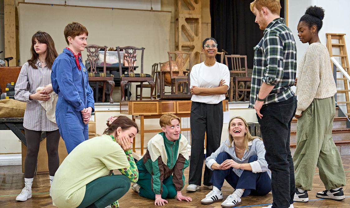 A group of actors are stood and sat together in a rehearal room. Three people are sat and kneeling on the floor in the centre looking up and smiling at a man in front ofthe, who looks down at them seriously. Others are stood around smiling.