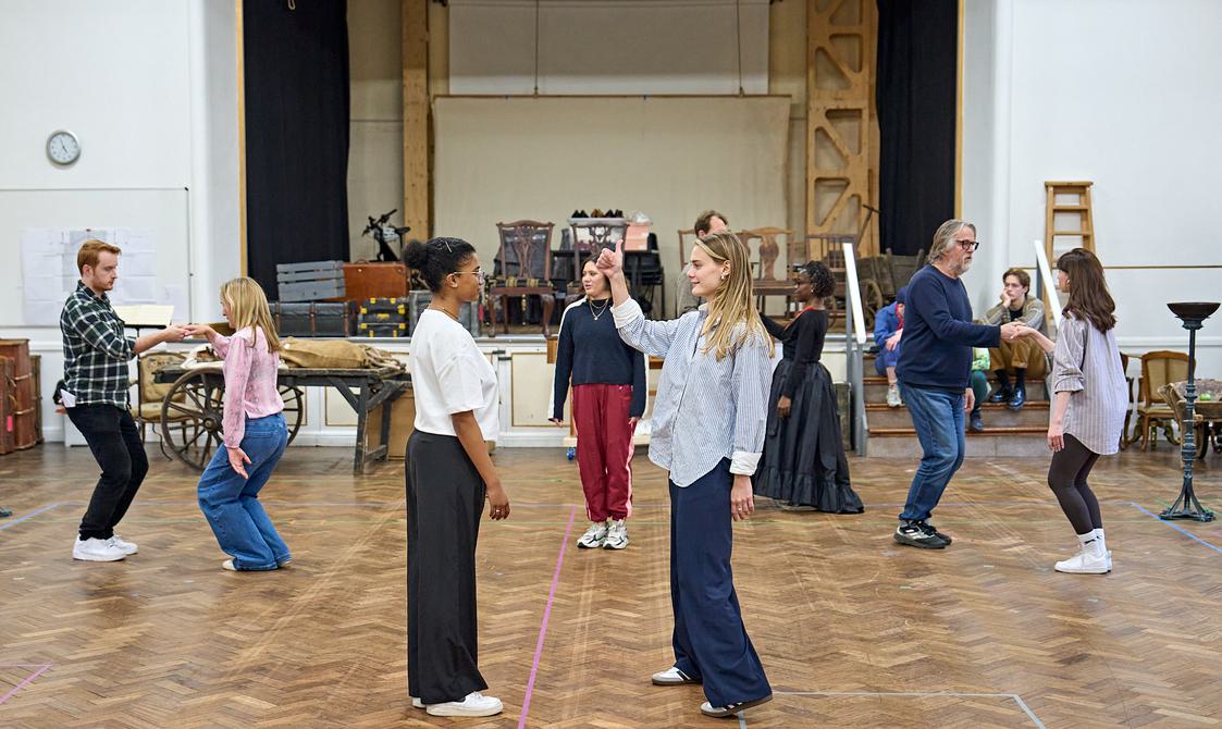 A group of actors stand in a rehearsal room, dancing in pairs. On each side are two pairs, holding each other's right hands, with their knees bent as if they are greeting one another. In the centre two women face each other, one has her right arm in the air giving a thumbs up.