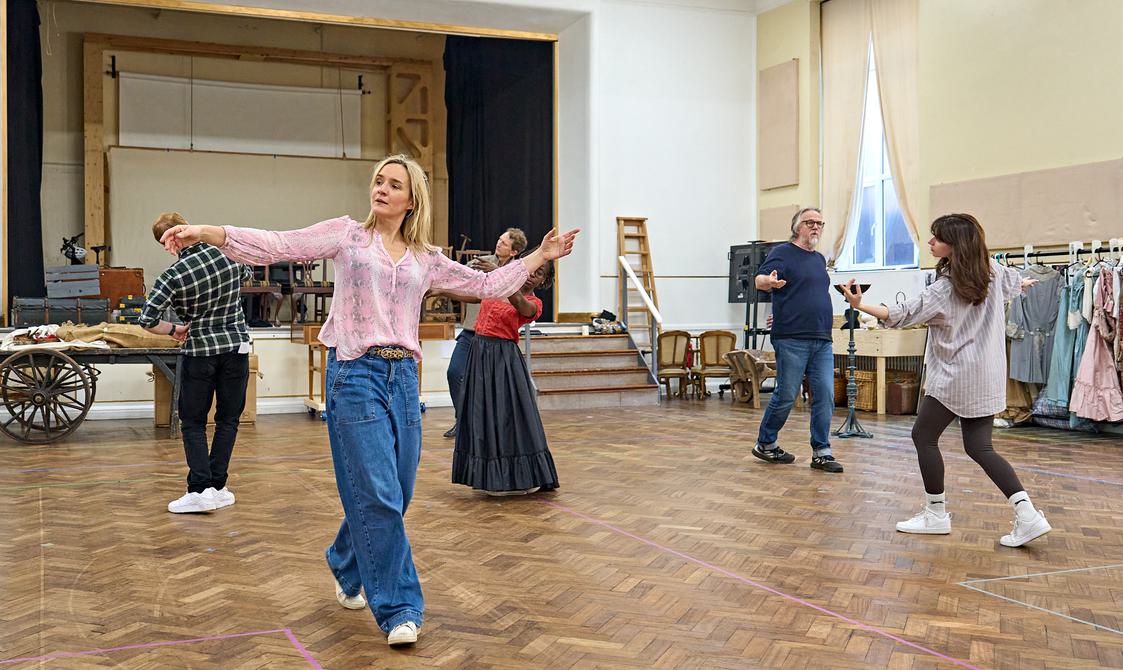 A group of actors are spread out in a rehearsal room. They are in stately poses, as if they have been slow dancing on their own, all facing in different directions. The floor is wooden and in the background there is a costume rail and a cart with big wooden wheels.