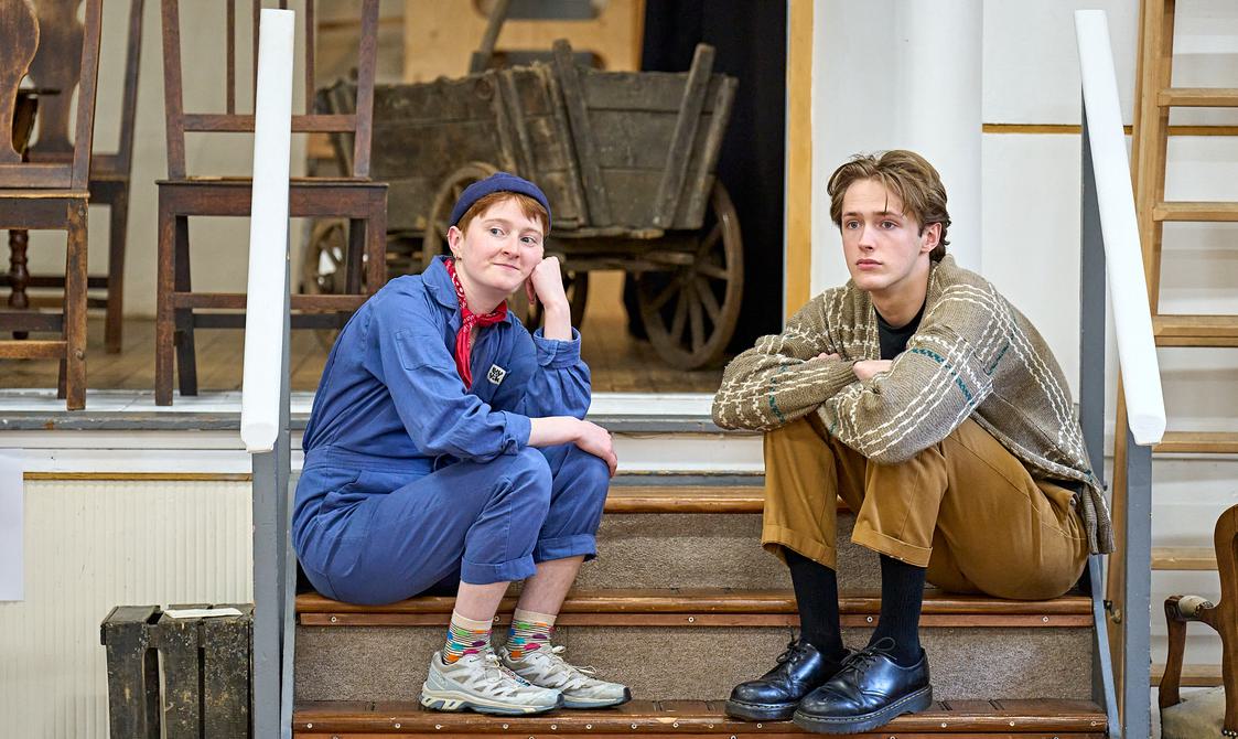 Two young actors sit on a set of steps, they look like they are wating for something. The person on the left is wearing blue overalls and white trainers, and rests their chin on their arm with a slight smile.. The person on the right is wearing brown trousers and a chequered shirt. Their arms are crossed as they stare into the distance.