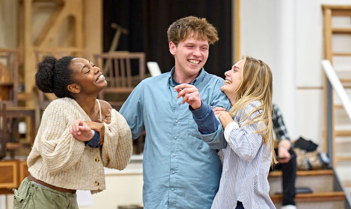 In the centre of the picture is a young blond man, wearing a blue shirt and blue jeans, smiling. Two young women are on either side of him, holding onto his arms and smiling and laughing as they look up at him. The woman on the left wears a cream cardigan and green cargo trousers, the woman on the right a blue and white shirt.