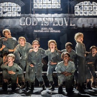 A group of children wearing dirty grey clothes are standing and crouched over holding their tummies as they sing.