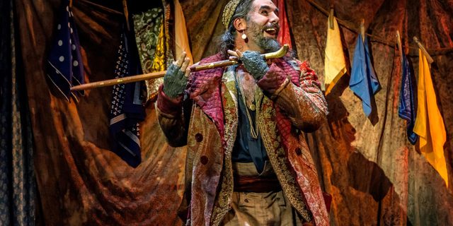 A man playing a walking stick as though it were a flute is wearing a colourful long coat, open blue shirt, fingerless gloves and jewellery. He has long greying hair and is wearing a cap. Behind him is a backdrop made out of scarves of different colours pegged up onto curtains.