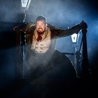 A man grimacing and bearing his teeth is holding on to the railings on a bridge. He has short dark hair and is wearing a long dark coat, light checked waistcoat and red tie around his neck.