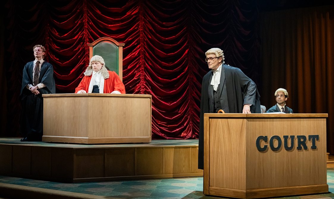 A scene in a courtroom. Towards the back on the left, a judge in wig and red robes sits behind a wooden desk on a tall-backed green leather chair, with a legal clerk to his right. On the right of the picture stands a barrister in legal robes and wig, wearing glasses and addressing someone out of shot to the left. Seated next to him is a younger barrister, also in a wig, behind a desk on which the word 'Court' is emblazoned.