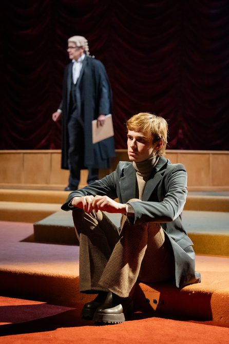 A young fair-haired man sits on a low carpeted step, resting his arms on his knees and with a thoughtful expression on his face. He wears a grey velvet jacket, brown rollneck and brown checked trousers. Behind him, out of focus, stands a man in barrister's gown and wig, carrying a brown paper file.