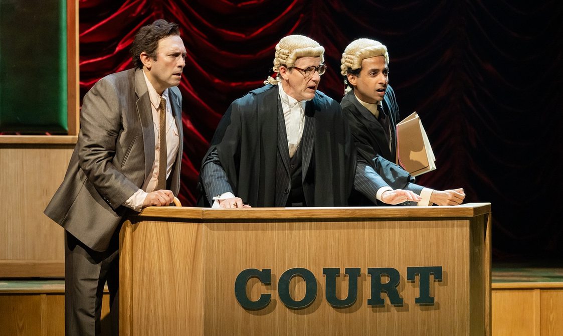 Three men stand behind a wooden desk on which is emblazoned the word 'Court', staring out to their left with shocked expressions. On the left, the man wears a shiny brown suit and tie; the other two men both wear barristers' legal dress and wigs. The older man in the centre wears glasses, and the younger man on the right carried a sheaf of papers under his arm.