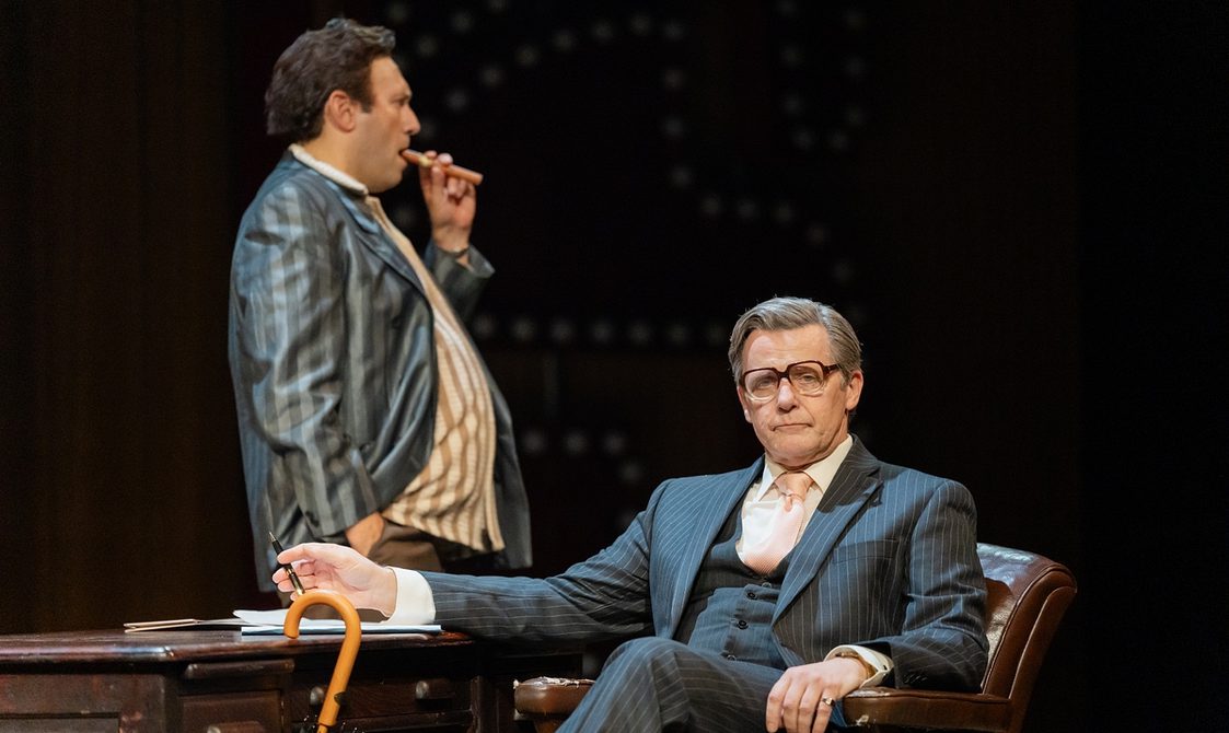 A middle-aged man with grey hair and glasses, wearing a dark three-piece pin-stripe suit, sits at a desk and stares out at the camera with a serious, unamused expression. An umbrella is propped against the desk and he holds a fountain pen in his right hand. Behind the desk, a brown-haired man wearing a shiny grey striped suit stands staring to the right, smoking a large cigar.