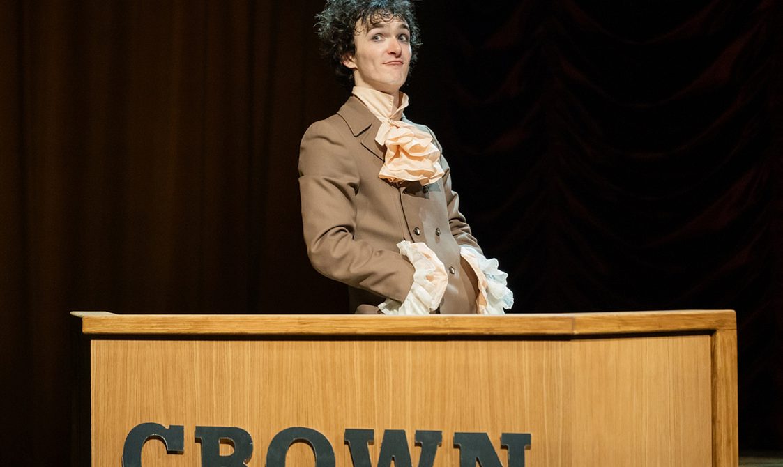 A young man with dark curly hair stands behind a wooden desk with the word 'Crown' emblazoned on it. He wears a Regency-style brown frock coat and cream shirt with extravagant ruffle at the neck and wrists, and looks out with a cheeky, irreverent expression.