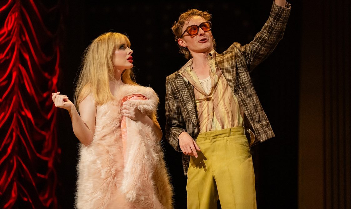 A young woman with long blonde hair stands with a large pink fur rug wrapped around her. Next to her is a man wearing sunglasses, with a brown check blazer, striped shirt and yellow trousers. They are both looking up to their left. A red ruched curtain hangs to the left of the picture.