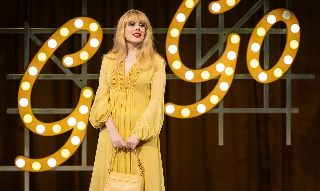 A young woman with long blonde hair and red lipstick stands smiling to her left, wearing a yellow boho dress with red embroidery and carrying a yellow handbag. Behind her are illuminated yellow letters saying Go-Go.