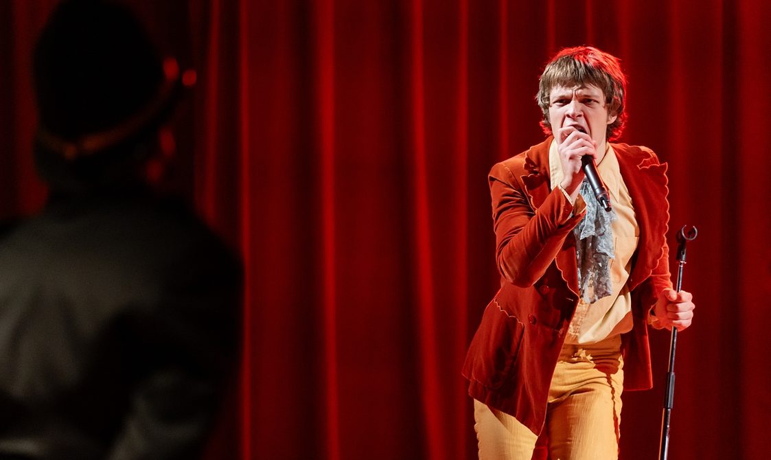 An actor playing Mick Jagger stands at the right of the photo, performing a song. He holds a microphone to his mouth, grasping the mic stand with his left hand, and wears 1960s style ochre coloured velvet jacket, yellow shirt and trousers with a grey cravat. Behind him is a dark red curtain and at left, out of focus, is the outline of a policeman.