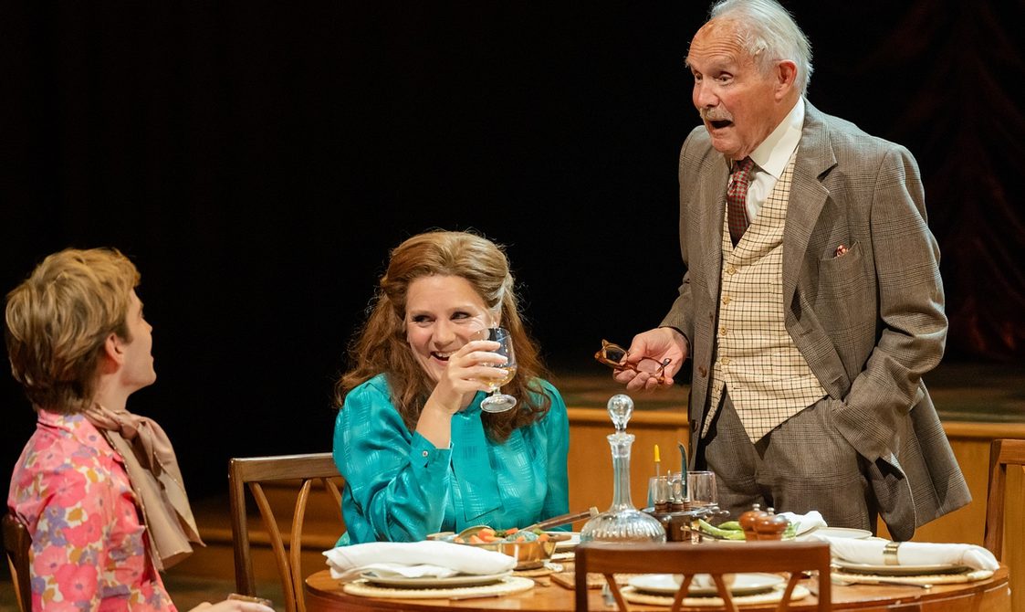 Three people around a table set for lunch. At left, a young man looks away from the camera to his companions, wearing a bright pink flowery shirt. In the centre, a woman with long brown hair swept back, sits smiling at the young man, wearing a turquoise blouse and lifting a wine glass. At right, an older man stands talking cheerfully to them as though telling a joke; he has white hair and a moustache and is smartly dressed in a brown suit, checked waistcoat and tie.
