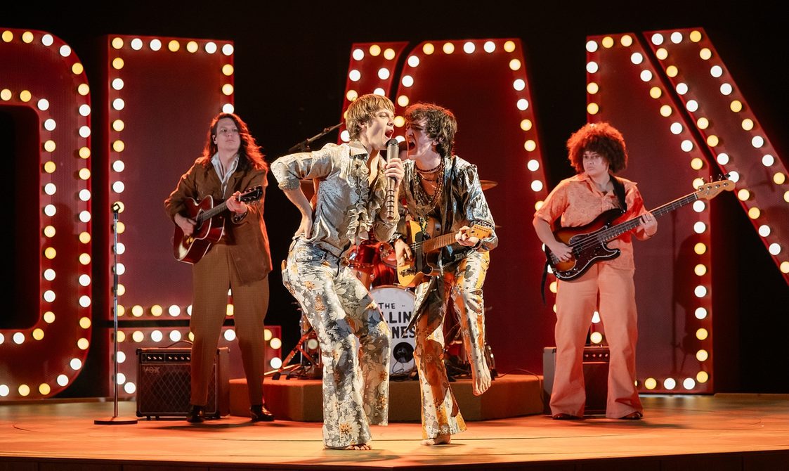 In the centre, two men in extravagant 1960s costumes - flowery flares and silvery satin shirts with ruffles - are giving an energetic performance. The man on the left is singing into a hand-held microphone with his right elbow bent and his hand on his hip. The musician on the right is also singing into his companion's microphone while playing a guitar. Behind them are two more guitarists and in the background are a number of large illuminated capital letters.
