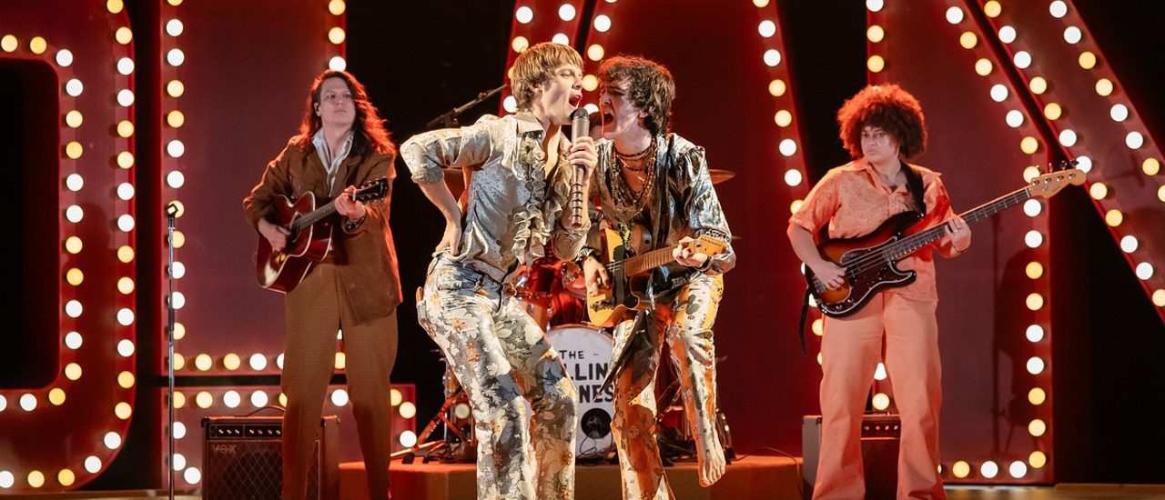In the centre, two men in extravagant 1960s costumes - flowery flares and silvery satin shirts with ruffles - are giving an energetic performance. The man on the left is singing into a hand-held microphone with his right elbow bent and his hand on his hip. The musician on the right is also singing into his companion's microphone while playing a guitar. Behind them are two more guitarists and in the background are a number of large illuminated capital letters.