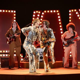 In the centre, two men in extravagant 1960s costumes - flowery flares and silvery satin shirts with ruffles - are giving an energetic performance. The man on the left is singing into a hand-held microphone with his right elbow bent and his hand on his hip. The musician on the right is also singing into his companion's microphone while playing a guitar. Behind them are two more guitarists and in the background are a number of large illuminated capital letters.