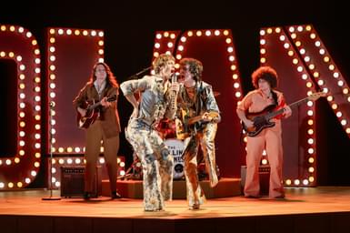 In the centre, two men in extravagant 1960s costumes - flowery flares and silvery satin shirts with ruffles - are giving an energetic performance. The man on the left is singing into a hand-held microphone with his right elbow bent and his hand on his hip. The musician on the right is also singing into his companion's microphone while playing a guitar. Behind them are two more guitarists and in the background are a number of large illuminated capital letters.