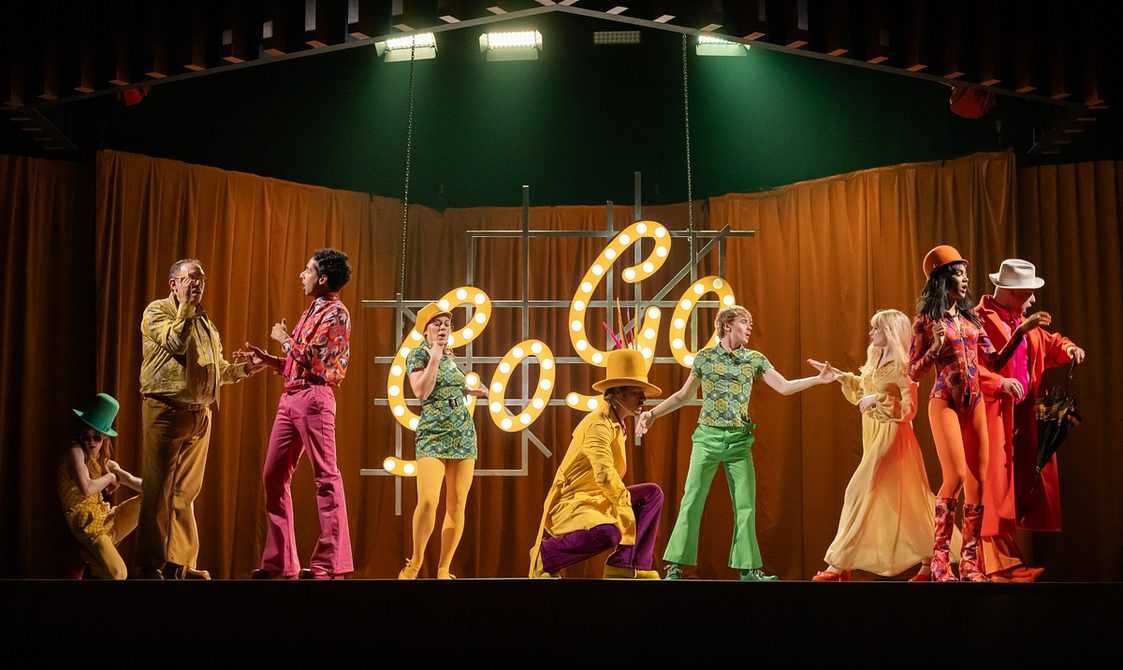 A group of people in brightly coloured 1960s-style clothes in shades of yellow, green, orange, purple and pink are dancing or posing against an orange curtain background, with the word 'Go-Go' spelled out in neon yellow lights.