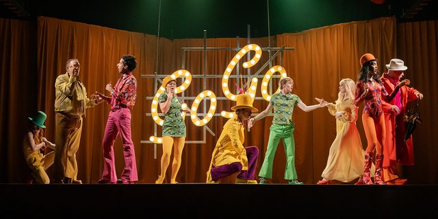 A group of people in brightly coloured 1960s-style clothes in shades of yellow, green, orange, purple and pink are dancing or posing against an orange curtain background, with the word 'Go-Go' spelled out in neon yellow lights.