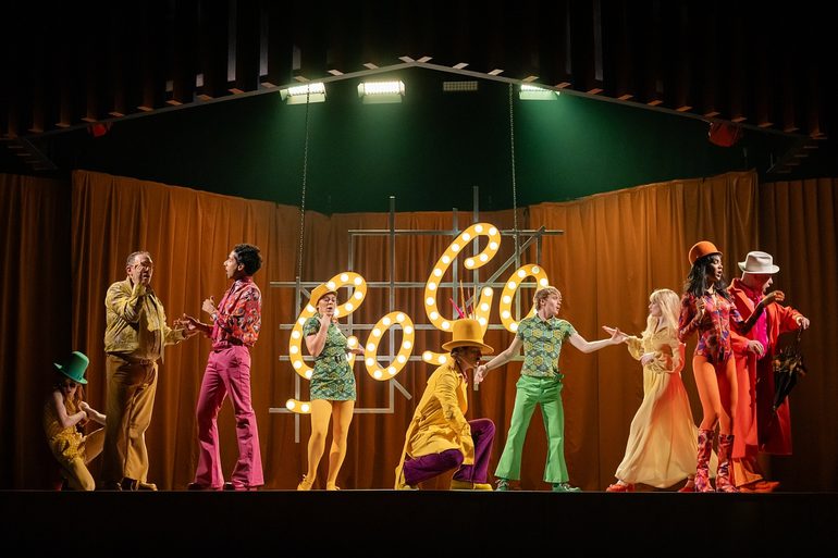 A group of people in brightly coloured 1960s-style clothes in shades of yellow, green, orange, purple and pink are dancing or posing against an orange curtain background, with the word 'Go-Go' spelled out in neon yellow lights.