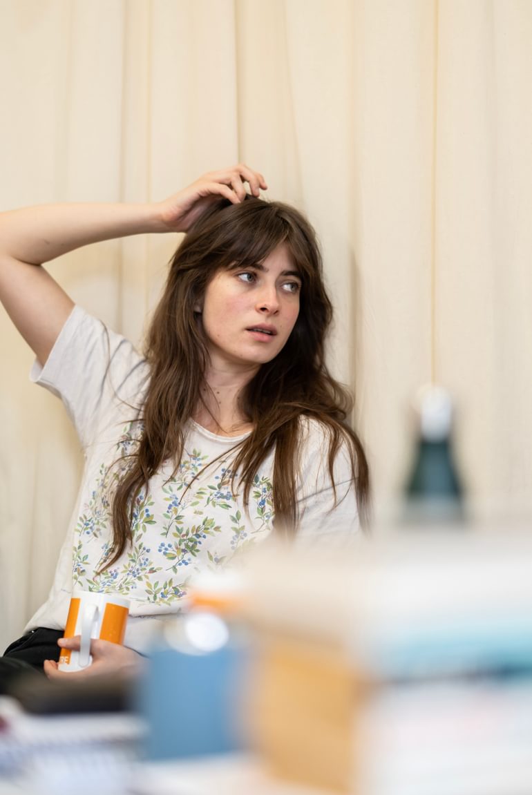 A woman with long dark hair sits looking to her left, her right arm raised to touch her head. She holds an orange mug in her left hand.