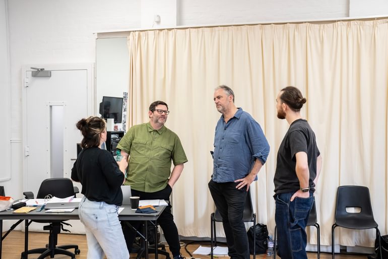 Four people stand in conversation with each other. Two look away from the camera towards the writer David Eldridge, wearing a green shirt and glasses, and director Jeremy Herrin, in a blue shirt, who are facing out.