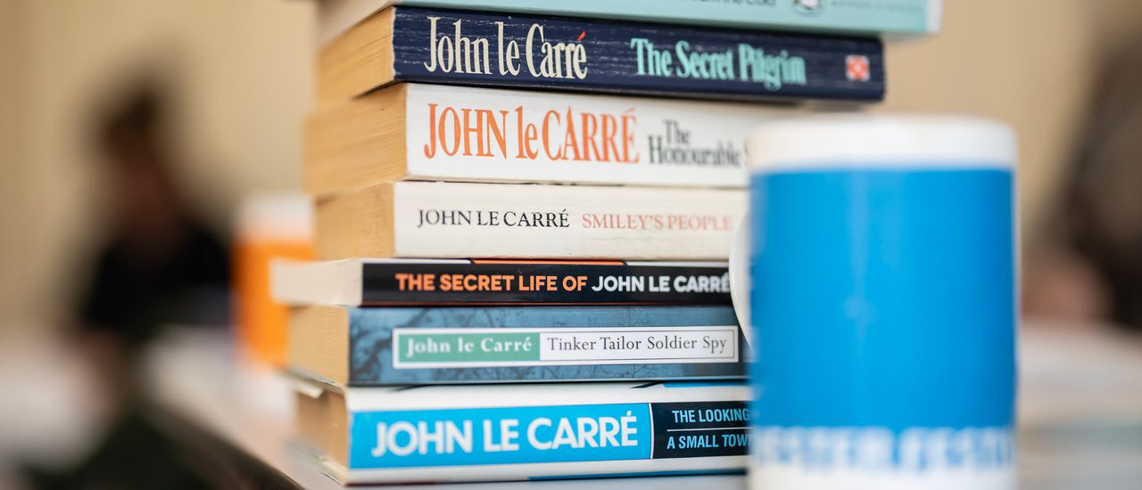 A pile of books by and about John le Carré on a table, behind a blue mug.