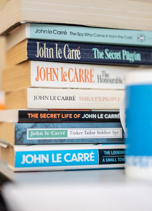 A pile of books by and about John le Carré on a table, behind a blue mug.