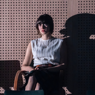 A young woman sits in a chair lit in moody lighting, so that her shadow appears oversized behind her. She looks off into the distance with a blank, absent expression.