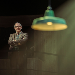 A man stands alone behind a handrailing. Behind him is an industrial, metal-clad set. In the top right corner is a bright spotlight, shining down. The man has a serious look as he folds his arms and stares down. He exudes authority and power.