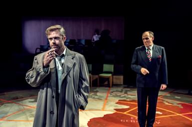 Two men stand on a stage that looks like a war map. The man in the foreground wears a long trench coat and smokes a cigarette. The man in the midground wears a smart suit and striped tie.