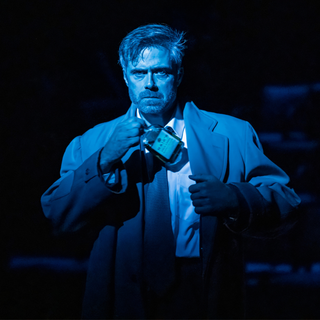 A man in a smart suit and tie is standing alone on the stage, dramatically lit in blue tones. He stares straight ahead with an intense, enraged stare, putting something into the pocket of his jacket.
