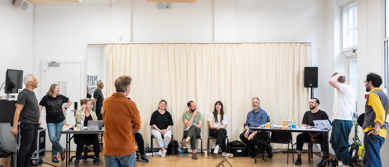 A group of people stand and sit in a rehearsal room, laughing and smiling at each other. A man in an orange shirt and jeans stands in centre with his back to us; to his right, an overturned chair lies on the floor.