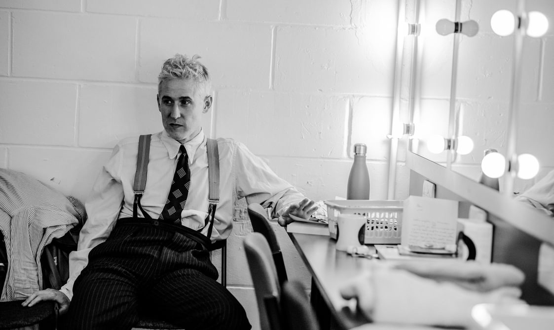 A black and white photo of a performer sitting in his dressing room, leaning one hand on the dressing table on the right and the other on the chair he is sitting on. He looks off to the side with an amused expression. He is wearing pin striped trousers, a white shirt, braces and a tie. His light hair is pushed back off his face. There are a variety of items spread across the dressing table on the right and mirrors surrounded by lights above it.
