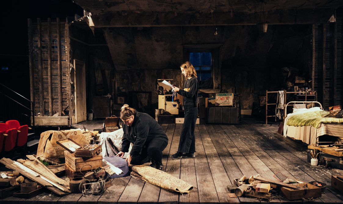 Two members of the crew dressed in black are on stage, dressed with the set for The Caretaker. One of them stands centre stage, reading from a clipboard, and the other bends down, opening a suitcase and looking down at it. They are both concentrating. The floor is covered with distressed, dark wooden floor boards. The walls and slanting ceiling are covered with peeling wallpaper and there are distressed wooden panels on each side. There is a cream door on the right hand side and a window in the centre, covered with a sagging piece of old sacking. The stage is covered with various junk and random objects, all from the 1950s, including rugs, suitcases, wooden boxes, tools and a white metal bed dressed with a red and cream stripey blanket and green satin quilt.