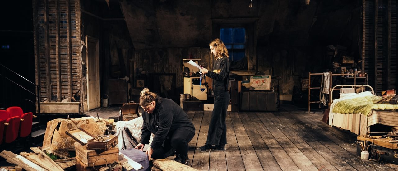 Two members of the crew dressed in black are on stage, dressed with the set for The Caretaker. One of them stands centre stage, reading from a clipboard, and the other bends down, opening a suitcase and looking down at it. They are both concentrating. The floor is covered with distressed, dark wooden floor boards. The walls and slanting ceiling are covered with peeling wallpaper and there are distressed wooden panels on each side. There is a cream door on the right hand side and a window in the centre, covered with a sagging piece of old sacking. The stage is covered with various junk and random objects, all from the 1950s, including rugs, suitcases, wooden boxes, tools and a white metal bed dressed with a red and cream stripey blanket and green satin quilt.