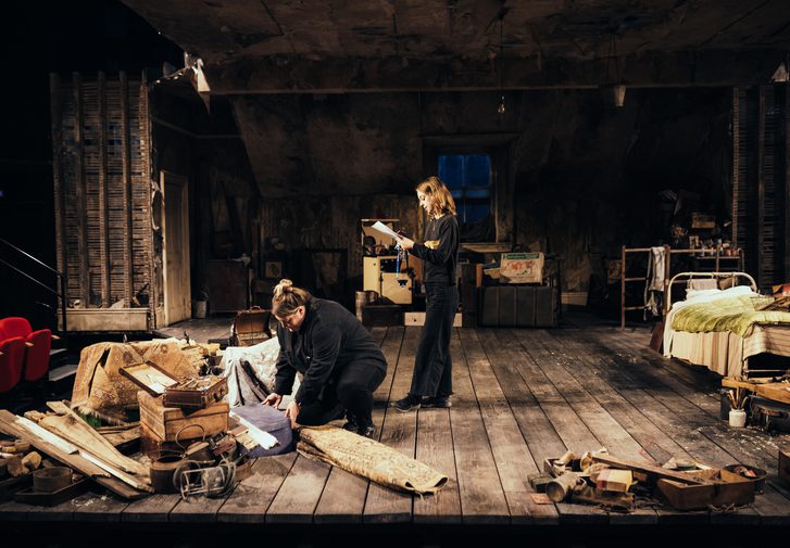 Two members of the crew dressed in black are on stage, dressed with the set for The Caretaker. One of them stands centre stage, reading from a clipboard, and the other bends down, opening a suitcase and looking down at it. They are both concentrating. The floor is covered with distressed, dark wooden floor boards. The walls and slanting ceiling are covered with peeling wallpaper and there are distressed wooden panels on each side. There is a cream door on the right hand side and a window in the centre, covered with a sagging piece of old sacking. The stage is covered with various junk and random objects, all from the 1950s, including rugs, suitcases, wooden boxes, tools and a white metal bed dressed with a red and cream stripey blanket and green satin quilt.