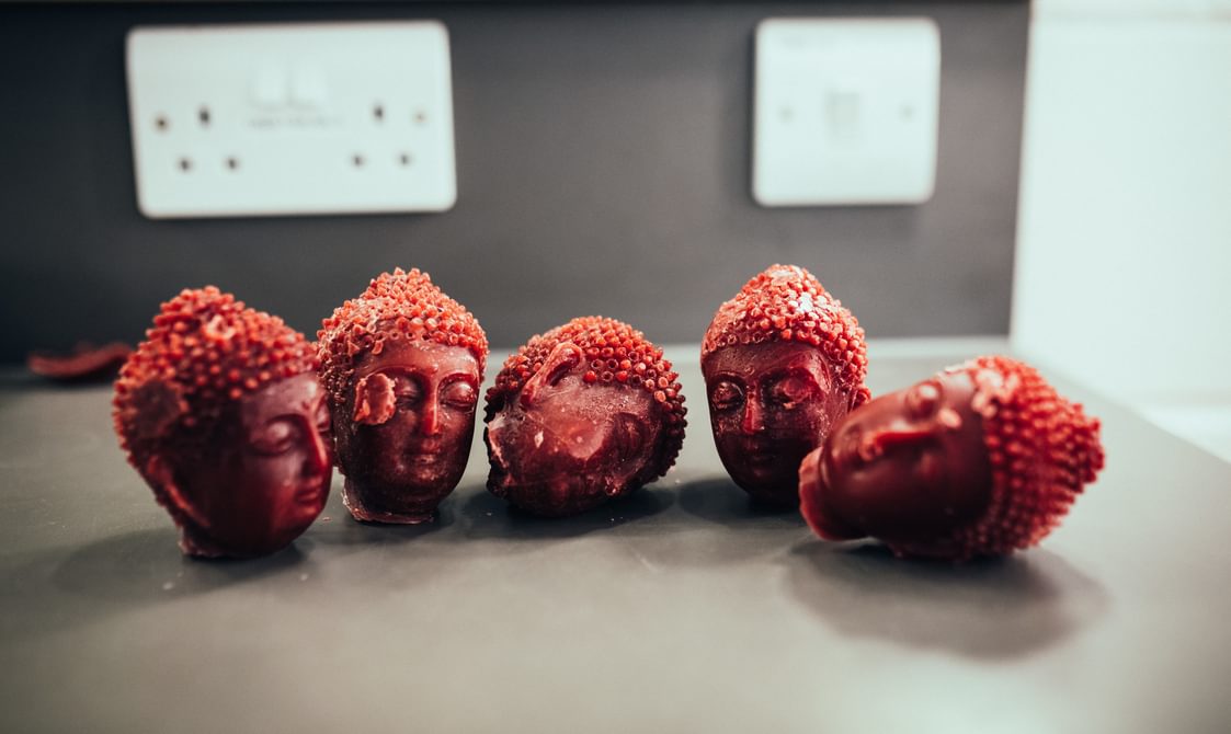 Five wax buddha heads sit on a grey desk, with a white plug socket out of focus in the background. Two of the heads have rolled onto their sides and they have all been damaged slightly - chipped or slightly broken.