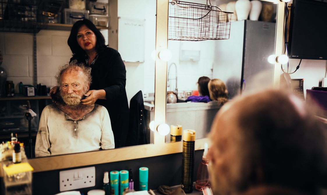 An elderly actor is photographed from behind sitting in front of a dressing room mirror, so his reflection can be seen. He is looking at his reflection with a serious, pensive expression. Behind him, a lady dressed in black with dark hair and red lipstick is carefully spreading black make up over his face. His face, head, neck and costume are smeared with it, so he looks dirty and tired. Various cosmetics can be seen on the dressing room underneath the mirror, with wigs and storage boxes in the background around the room.
