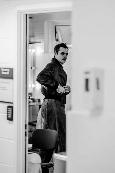 A black and white photo of a performer in his dressing room, taken from outside the door. He is looking behind him with an intense expression, putting something in his pocket. He is wearing grey trousers and a dark leather jacket.