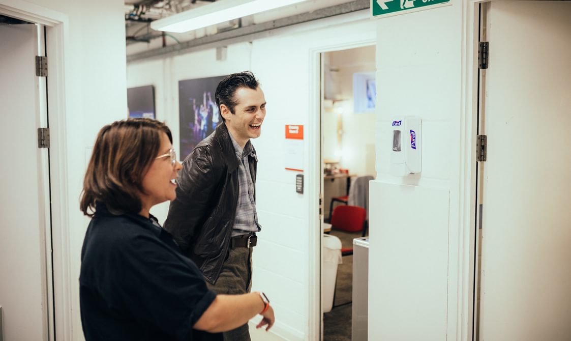 A member of the crew and a performer photographed side on in a hallway. The walls are white and a door is open to reveal part of a dressing room with a desk, mirrors and a red chair. The lady on the left is wearing a black shirt and talking. The man on the right is wearing grey trousers, a dark belt, a blue checked shirt and a dark leather jacket. His dark hair is slicked back and he is laughing. They are both looking off to the right as if talking to someone.