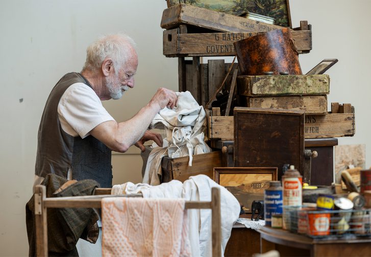 Actor Ian McDiarmid stands next to a tall pile of wooden crates, paintings and boxes in a rehearsal room. He is looking through a messy pile of white clothes or bedding. In front of him is a clothes drying rack and a table covered in paint and spray cans.